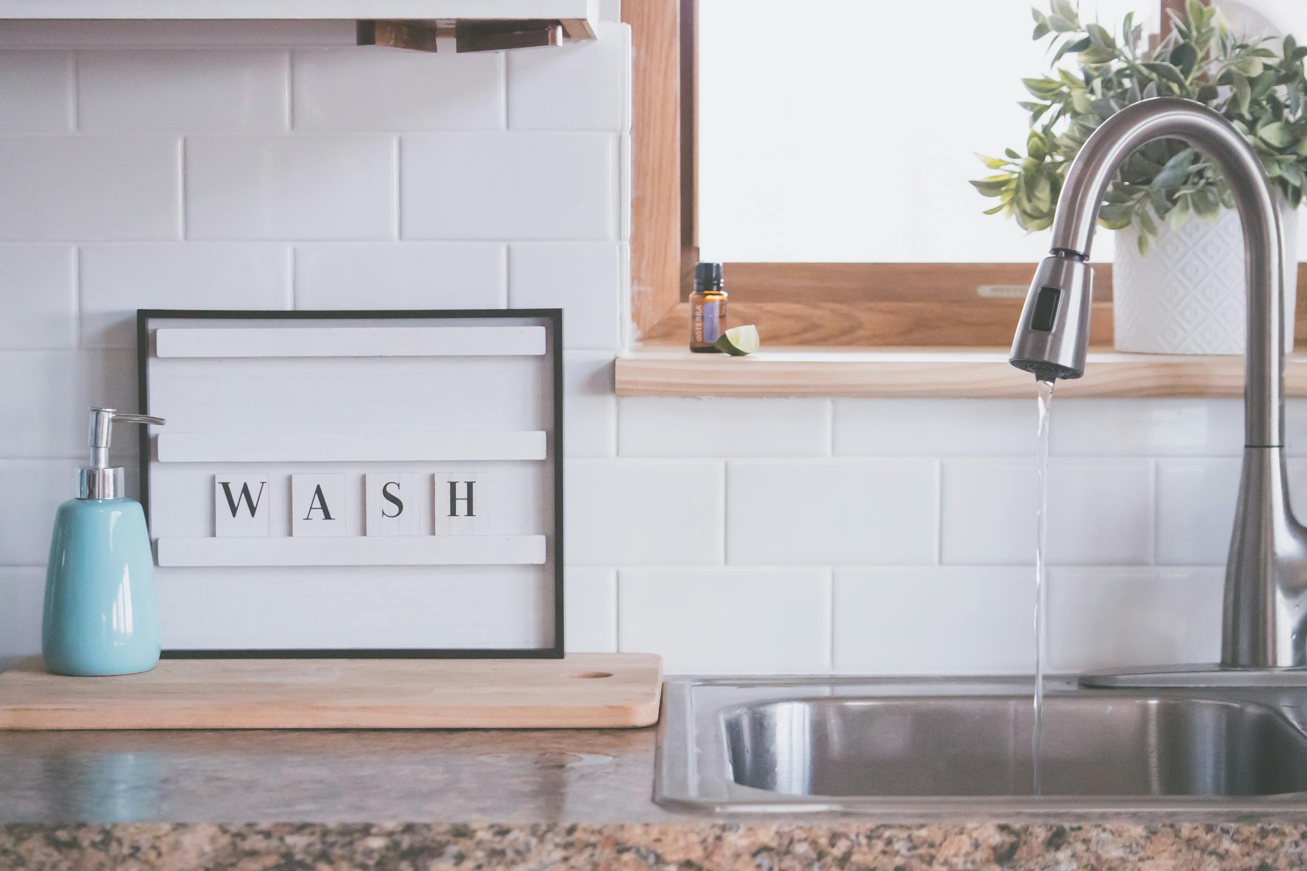 Board with word WASH placed near soap dispenser on counter in bathroom with water streaming from open faucet